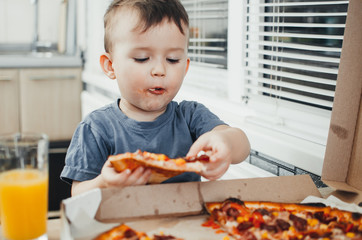 the little boy eats a huge harmful pizza himself in the kitchen and drinks juice, very fat and...