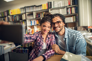 Close up of smiling hipster cheerful young student love couple sitting in the high school library and taking a selfie near the sunny window.