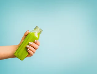 Fotobehang Female hand holding bottle with green summer beverage: smoothie or juice at blue background. © VICUSCHKA