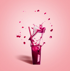 Glass with drinking  straw and purple splash summer beverage: smoothie or juice on pink background, front view