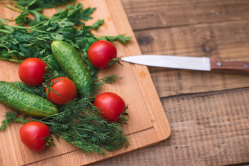 Cherry tomatoes, cucumbers, dill and parsley on wooden board on table with knife closeup. Healthy food concept.