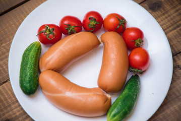 Cherry tomatoes, cucumbers and raw sausages on white plate on wooden table closeup