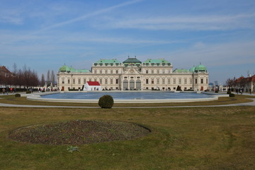 Piękny pałac w Wiedniu, na pierwszym planie trawnik z okrągłym, przystrzyżonym zielonym krzewem, kamienna sadzawka, niebo błękitne z pojedynczymi malowniczymi pasmami chmur