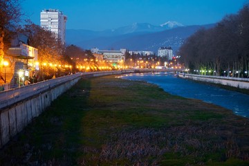 River valley in the city center. Green grass and blue waters of the river. Lights of the evening city.