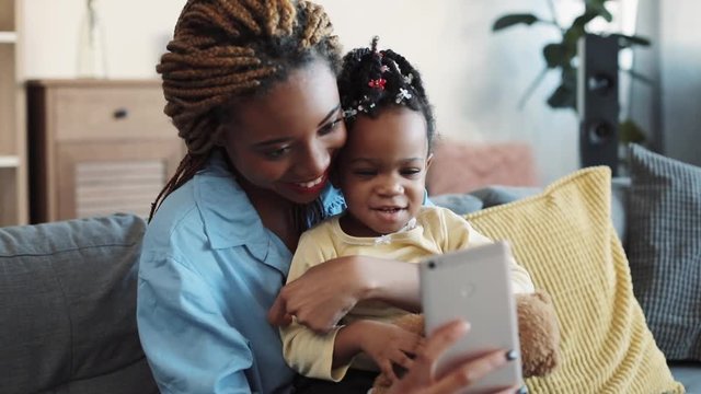 Beautiful stylish young mother with dreadlocks takes selfie with her cute smiling daughter and teddy bear at home. Modern culture, family portrait. Slow motion, close up view