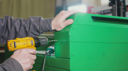 Worker man using an electric hand drill - making hole in green metal machine