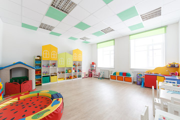 Spacious white game room in the kindergarten with toys, two large windows and tables for classes.