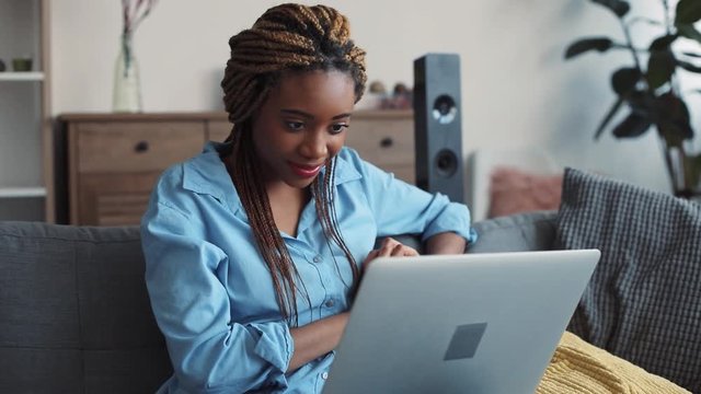 Pretty young stylish woman with dreadlocks sits on the sofa with many cushions in the cozy living room, and surfs the internet. Blogger, modern lifestyle. Slow motion, close up view