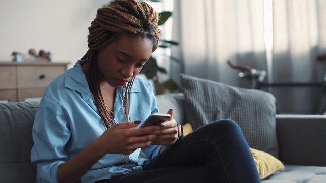 Lovely young woman with dreadlocks smiles as she texts messages via her smartphone while relaxing on the sofa in her cozy living room. Close up view, female portrait