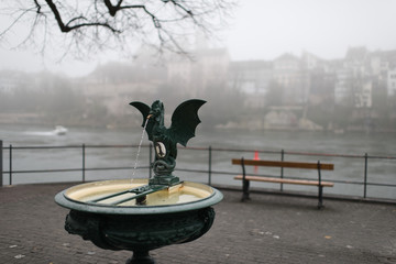 city fountain basel switzerland, foggy day with rhine river and old town in background - obrazy, fototapety, plakaty