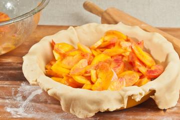 Top view of ripe, raw sliced peaches on rolled out dough in a ceramic pie plate with a wood rolling pin on a wood cutting board with dusting flour, peach pie