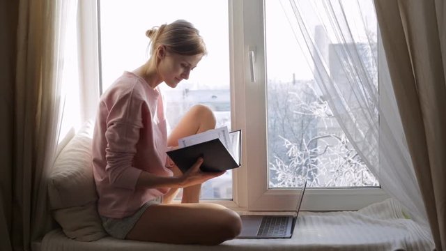 Beautiful young woman with a notepad sitting on window sill. Winter outside