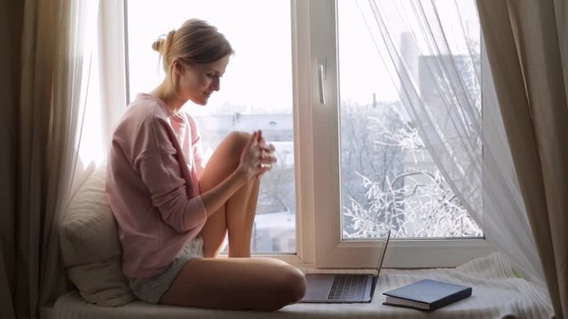 Young woman with laptop sitting on windowsill. Then she looking through the window.