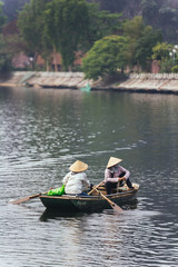 Fototapeta na wymiar Rowing boat women wearing conical hat sitting in and rowing a boat with paddles over the river in background at Trang An Grottoes in Ninh Binh, Vietnam.