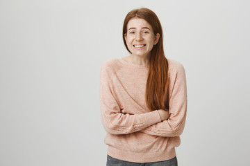 Indoor shot of kind charming ginger girl standing crossed hands as if she is freezing, smiling awkwardly and looking at camera over gray background. Woman went outdoor without wearing jacket