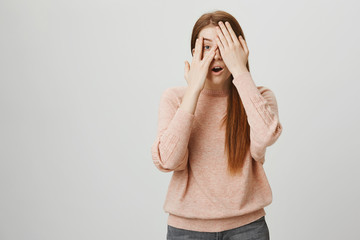 Attractive european redhead woman covering eyes with hands and peeking through it, being curious what will happen, standing over gray background. Girl is scared of horror but also very interested.