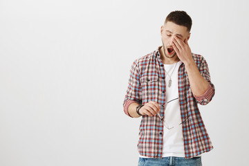 Man came from nightshift and needs some sleep. Portrait of cute unshaven male student yawning and rubbing eyes, being exhausted after work, holding glasses in hand. Guy is tired of doing homework