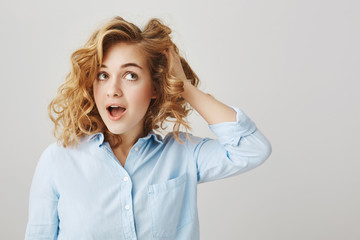 My hairdresser is fantastic. Studio shot of cute feminine caucasian girl with curly hair touching it and looking aside with opened mouth, expressing sensuality while being over gray background