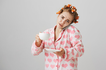 Portrait of bothered funny young woman, being busy with talking on smartphone and trying to finish coffee, wearing hair curlers and pajamas with hearts. Girl is late on work and tries to hurry up