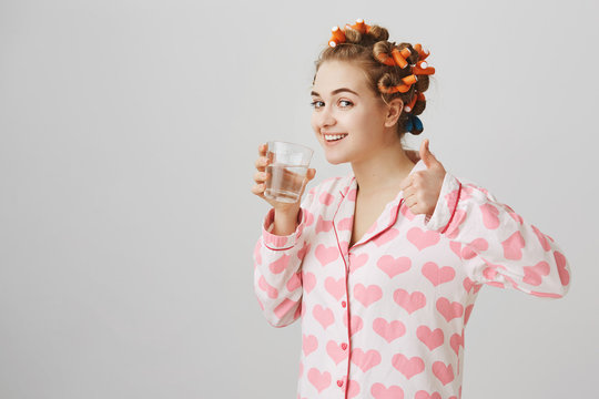 Funny attractive blonde wearing pajamas with hearts and hair curlers, standing half-turned, drinking water, smiling at camera and showing thumb up over gray background. Girl supports healthy lifestyle