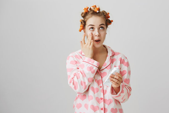 Cute feminine european girl wearing hair-curlers and pyjamas applying cream on clean skin, looking up and standing over gray background. Fashionable woman takes care of her appearance