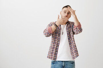 Shocked embarrassed attractive man in glasses trying to hide face behind palms, peeking through hands with surprised expression, standing against gray background. Shy guy saw naked woman on balcony