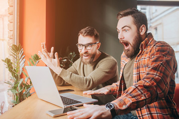 Very emotional picture of guys looking to the laptop's screen. One of them is very amazed and opened his mouth very wide while another is just looking to the screen.