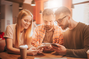 Nice picture of three friends sitting together and holding phones in their hands. They are playing games. This people are happy but concentrated.