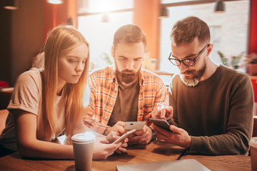 Good picture of three concentrated friends sitting together at the table and looking to their phones. They are playing games.