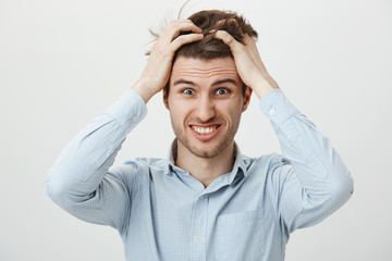 Portrait of funny young man scratching head, grimacing and smiling nervously, meeting deadline and trying to finish work in time, being bothered with time limits over gray background