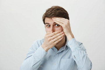 Is it over now. Portrait of scared and shocked young attractive man covering face with hands as if hiding or feeling fear, peeking at camera through hole, standing against gray background