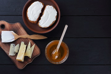 Cheese on a wooden board, sandwiches with soft cheese, honey in glass dishes, copy space, minimalism, cheese pattern, wooden dark background, healthy food, wooden cutlery, art