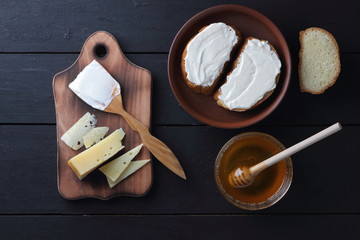 Cheese on a wooden board, sandwiches with soft cheese, honey in glass dishes, copy space, minimalism, cheese pattern, wooden dark background, healthy food, wooden cutlery, art
