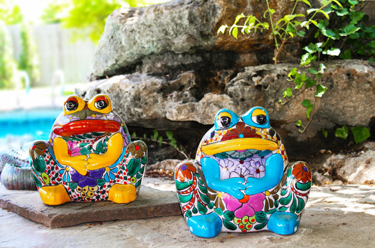Two brightly colored and decorated ceramic frogs sit by landscaping rocks by swimming pool