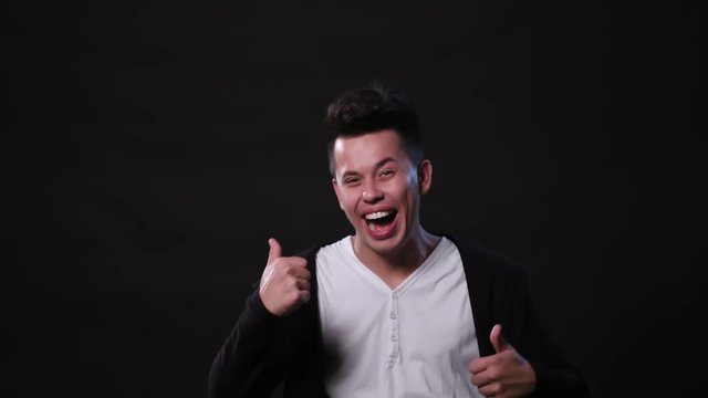 An attractive young man showing a Thumbs Up gesture against a black background. Medium Shot