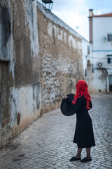 Fototapeta na wymiar A small witch in a black dress with red hair is holding a hat in her hands and standing with his back against the backdrop of the street of the old city
