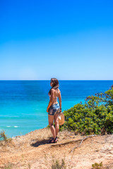 A girl with a hat in her hands is standing with her back on a rock against the blue ocean against the beach of Praia de Boião, Portimao, Alvor, Portugal.