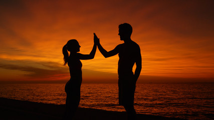SILHOUETTE: Young man and woman high five by the ocean at sunset after workout.