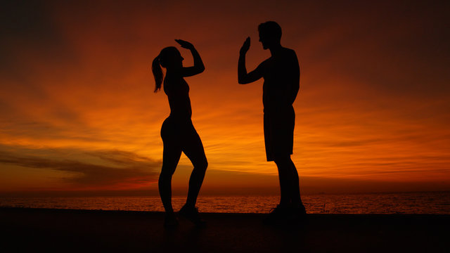 LOW ANGLE, SILHOUETTE: Happy young man and woman high five at stunning sunset.