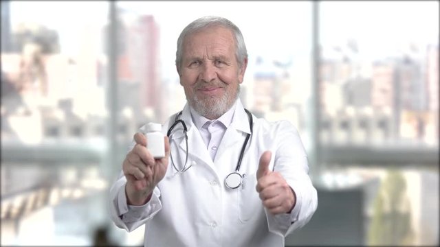 Elderly smiling doctor showing bottle with pills. Aged caucasian doctor holding container with pills and showing thumb up. Pharmaceutical and medicine concept.