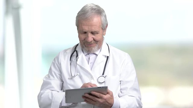 Elderly doctor typing on pc tablet. Senior male doctor using computer tablet on blurred background. People, profession, technology.