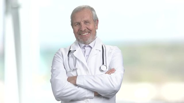 Cheerful grey haired doctor on blurred background. Portrait of happy senior doctor crossed arms and wearing medical equipment.