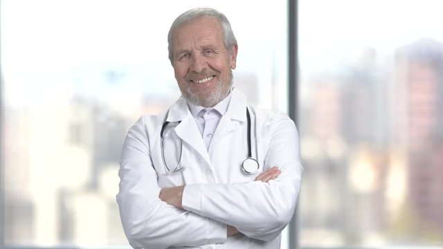 Senior handsome doctor with arms crossed. Happy older doctor smiling and looking at camera with folded arms, blurred background.