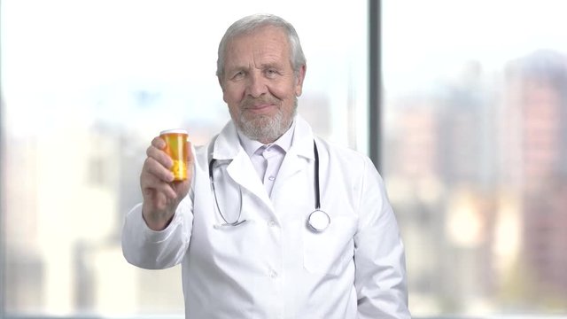 Cheerful doctor with pills, blurred background. Caucasian male doctor holding medication and showing thumb up gesture. New ways of therapy.
