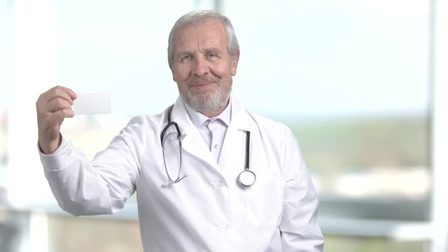 Senior doctor holding blank card for text. Cheerful male doctor showing paper card on blurred background. Card for contact information.