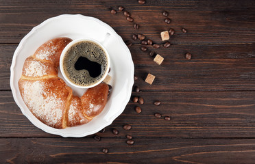 Plate with fresh tasty crescent roll and cup of coffee on wooden table