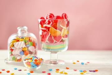 Glassware with sprinkles and different candies on table against color background