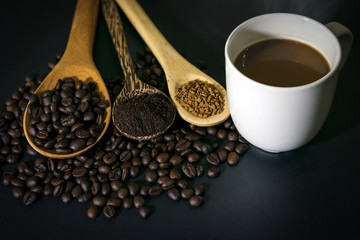 Hot coffee in white ceramic glass and coffee beans, coffee powder and instant coffee are placed in a wooden spoon placed on a black table top.