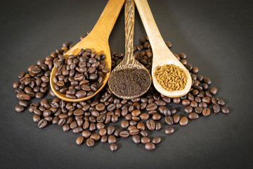 Coffee beans, coffee powder and instant coffee are placed in a wooden spoon placed on a black table top.