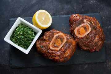 Stone slate tray with raw marinated cross cut veal shank and gremolata for cooking ossobuco, high angle view, studio shot